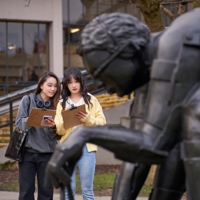 Two people holding clipboards and drawing a bronze sculpture of a man bent over in thought.