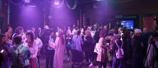 A crowd of people stand in a room with industrial features. They stand in smaller, intimate groups whilst talking and drinking. The lighting has a purple hue casting an atmospheric glow.  