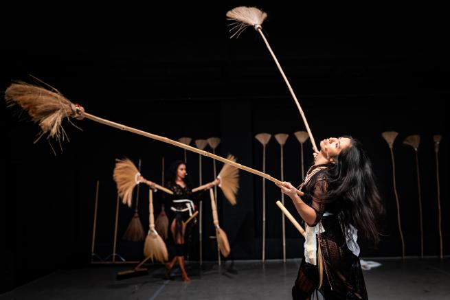 Two women, Eisa Jocson from the Philippines and Venuri Perera from Sri Lanka, perform under harsh spotlights, each holding long-handled straw brushes. Jocson stands in the foreground gazing upward, while Perera dances behind her.