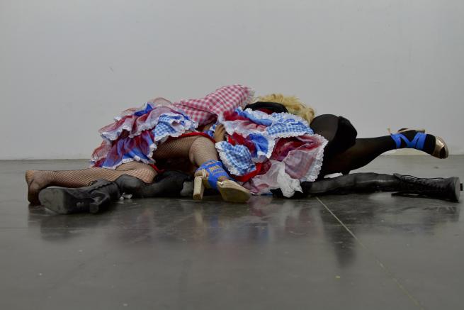 A group of people lie on a concrete floor, wearing blue, red, and white gingham dresses. Only their legs are visible, clad in tights and heeled shoes wrapped with blue ribbon.