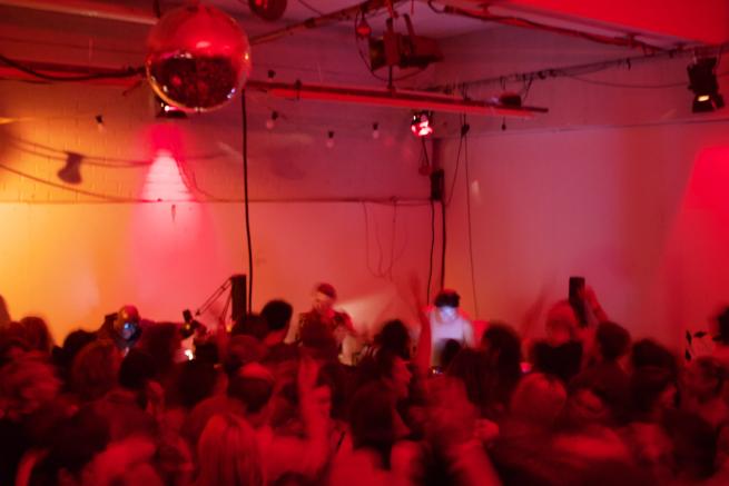 A group dances in a dimly lit room, bathed in red and orange light. A DJ, centered in the background with headphones on, plays records. The image is intentionally blurred to obscure faces.