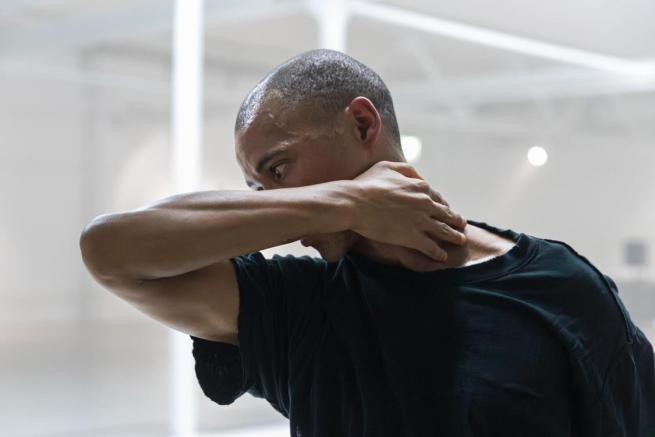 A black man is pictured in motion with his profile central to the image, he is visible only from the waist up. The room is brightly lit and has white industrial features. He has short, dark, afro hair and is wearing an oversized black t-shirt. His right arm extends out in front of him, bending at the elbow, his hand reaches around, resting on the nape of his neck.  