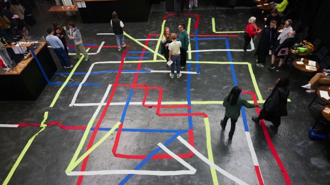A group of people are seen from above in a dimly lit room. Ribbons in red, green, white, and blue form a loose, overlapping grid on the floor.