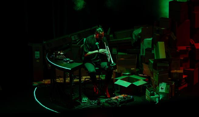 Ahamefule J. Olu sits centre stage surrounded by several objects including instruments, furniture and cardboard boxes. Wearing a black, short sleeve t-shirt and jeans they are capture in motion playing the trumpet. They have dark skin and cornrowed hair that reaches their shoulders. The room is in darkness yet Olu is visible through a soft, green spotlight. 