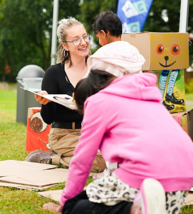 A woman sitting on grass reading a picture book to two young children at an outdoor event. She is smiling and holding the book open while one child sits in her lap and another leans in to look. Colorful cardboard boxes with painted faces are in the background