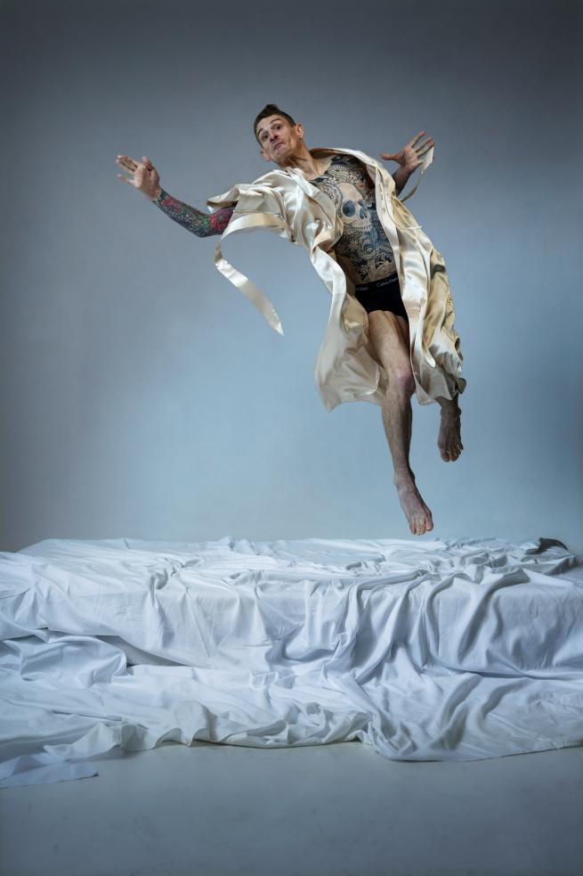 Dan Daw is captured in motion against a minimalist white backdrop. He is tall, white and crippled and his torso and arms are decorated in colourful tattoos. Wearing a cream silk robe and small black underpants, he is air-born as if he was floating. Underneath him sits a mattress, covered in a white, textured sheet.  