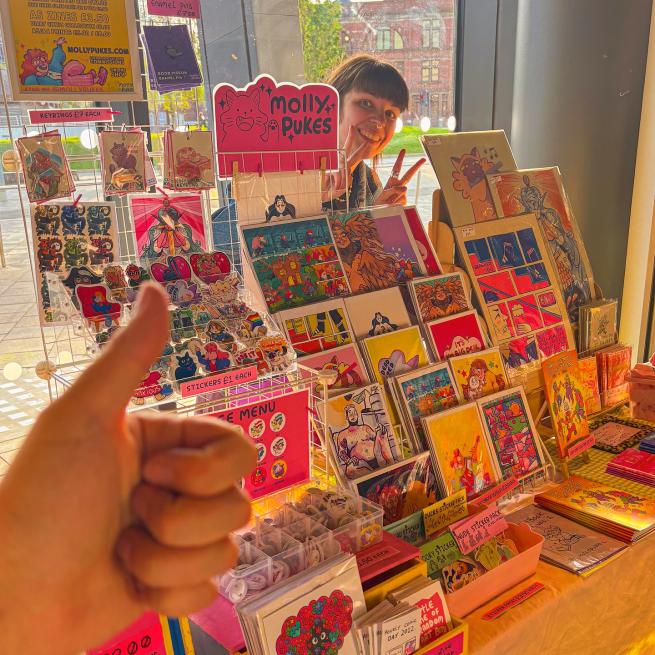 A local independant artist peeks out from behind their colourful stall full of comics, prints, and stickers as a hand in the foreground gives a thumbs up of approval