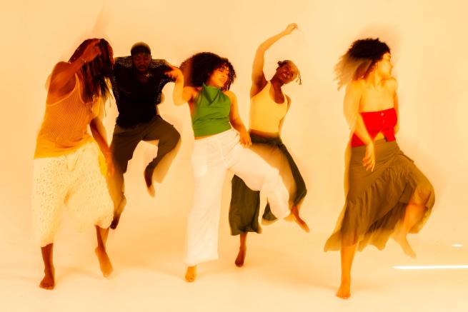 A group of dancers of mixed heritage are pictured in motion, against a bright yellow backdrop. The dancers each lift their left legs in the air whilst their right leg remains on the ground. They are each wearing loose fitting clothing in a range of brightly coloured earth tones. The image is intentionally blurred to emphasize the dancers movements. 