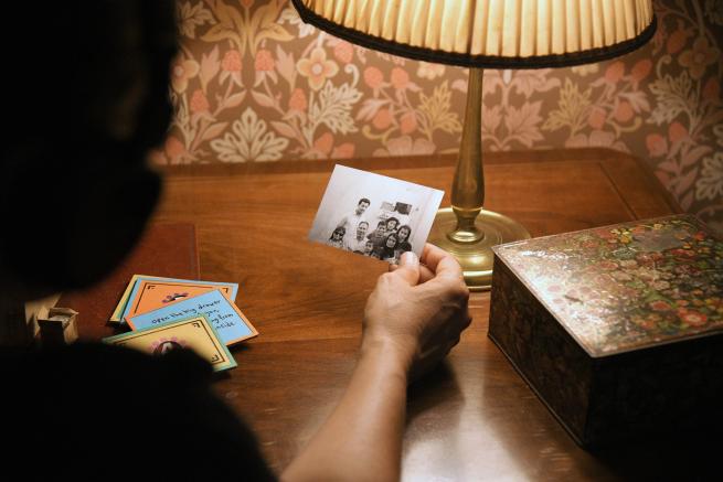  A Palestinian man sits with his back to the camera, leaning forward against a wooden desk. With only his arm is visible, as he sits in darkness, he is holding a black and white photograph comprising of several people of mixed ages and genders.    The desk rests against an wall covered in ornate pink and cream wallpaper. On top of the desk sits a metal lamp and a decorative wooden box. 