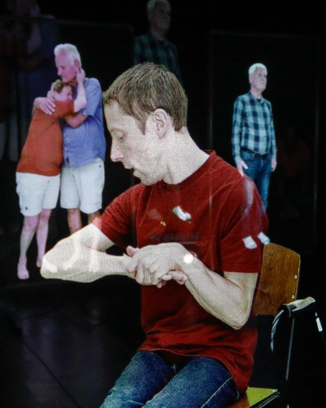 Central to the image sits a white man. As he sits on a wooden chair, he gazes sideways with his arms bent in front of him. He has short, light brown hair and is wearing a red, short sleeve t-shirt and blue jeans.    In the background stand 3 other people. Two are embraced, holding one another by the neck. The man is wearing a blue t-shirt and the woman is wearing a red shirt, both are wearing white shorts. To his right is a white man, he is wearing a blue check shirt and dark blue jeans.