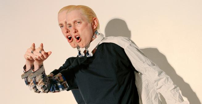 Pictured in motion, stands Eve Stainton against a cream backdrop. They are white with bright blonde scraped back hair, reaching just below their ears. Their shirt, in neutral, contrasting tones is a mixture of textures and materials. Their stance is strong, leaning exaggeratedly to the left. Their hand reaches forward as their index finger points out. Their image is duplicated and overlayed. 