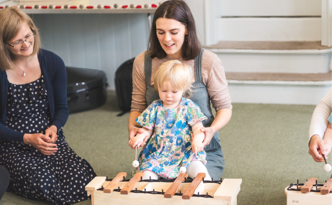 Music at Heart picture with teachers Caitlin and Carolyn supporting a child to play the xylophone