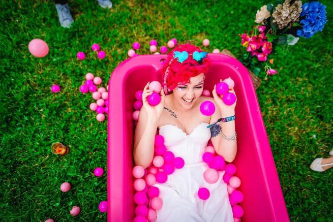 female presenting bride with pink hair in a white outfit, in a pink bath with pink balls 