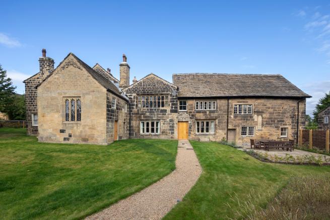  A wide, outdoor shot shows Calverley Old Hall, a historic stone building with a large green lawn in front of it. A light-colored gravel path extends from the foreground to a wooden front door in the center of the building. The hall is a sprawling structure with multiple sections and varying rooflines, all made of dark grey stone with many windows. To the right of the path, a patio with a large wooden table and benches is visible. The sky is clear and blue.