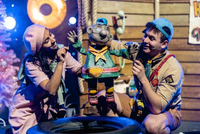 Two actors dressed as children hold a puppet of a third child character