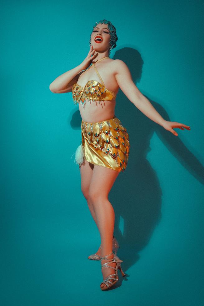 Picture of Aurora Nova, a showgirl wearing an embellished yellow costume in front of a turquoise backdrop