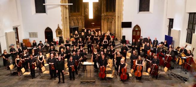 Full orchestra standing with their instruments wearing black clothes.