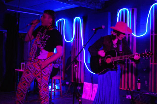 A photo of ThemTheres Cowboys performing at wharf. Lewis is in their dungarees with a colourful pattern and Spike is wearing a pinkc owboy hat, a blue skirt and carrying their guitar.