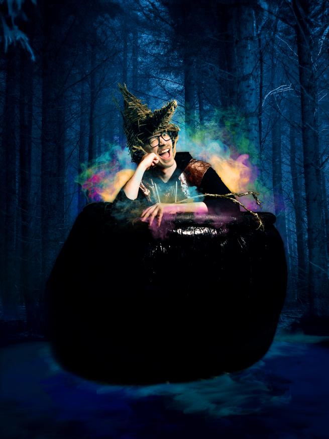 A person floats in a dark woods, surrounded by colourful smoke, with a witches hat on, and winks a the camera