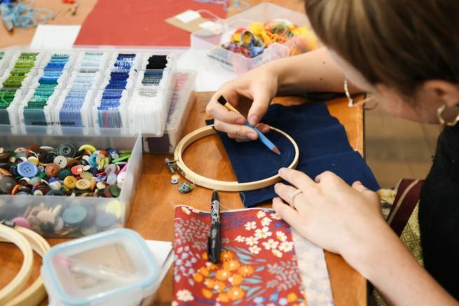 A person at a table drawing onto fabric by a hoop surrounded by sewing threads, fabrics and buttons.