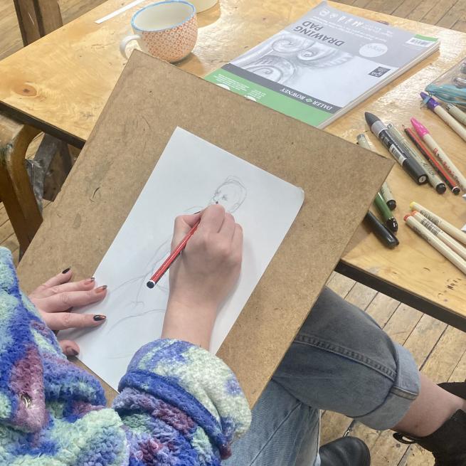 A photo taken over someones shoulder showing them holding a pencil over a sketch of a person