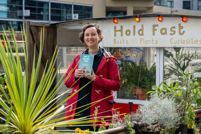 A picture of the author SJ Bradley on a boat (the bookshop Hold Fast) surrounded by plants. She has a red coat on and is smiling and holding a book (her work 'Maps of Imaginary Towns')