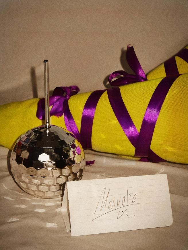 Legs wearing yellow stockings, and purple cross garters lay behind a disco ball cup and a letter addressed to Malvolio