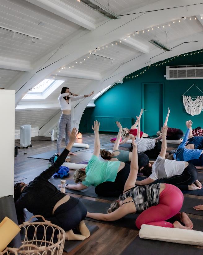 A yoga teacher guides a class in a shoulder stretch in a studio with fairy lights and arched ceiling.