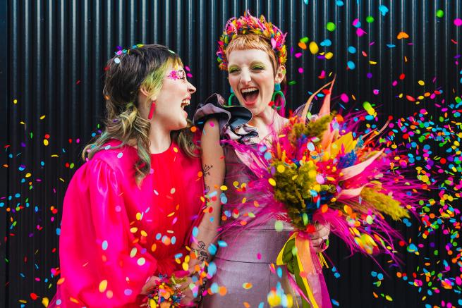 Two brides in colourful clothes with a colourful bouquet and confetti falling around them.