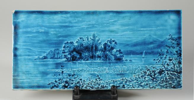 A blue ceramic tile featuring a scene of a river and vegetation and mountains.