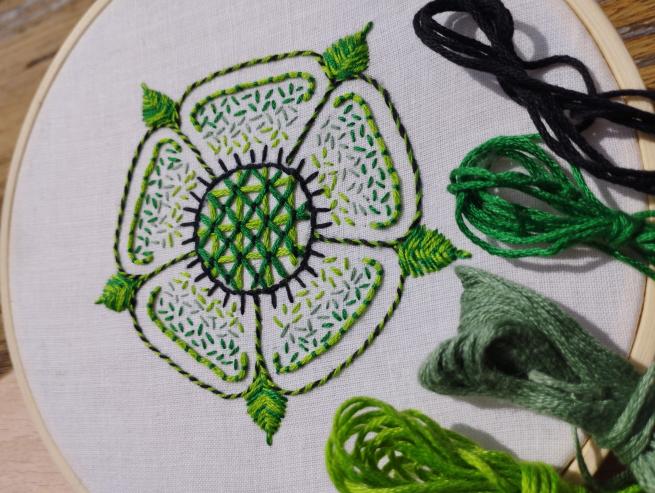  A close-up shot of a white fabric stretched on an embroidery hoop shows a Tudor rose design. The rose has five white petals, each filled with green stitches that create a textured, leafy pattern. The center of the rose is densely stitched in shades of green, forming a geometric, criss-cross pattern. The rose is outlined with a thin black and green border. To the right of the embroidery, loose bundles of green and black thread are visible.