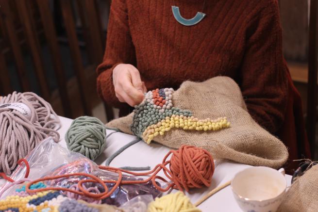  A person in a rust-coloured sweater works on a colourful rag rug, surrounded by balls of yarn.