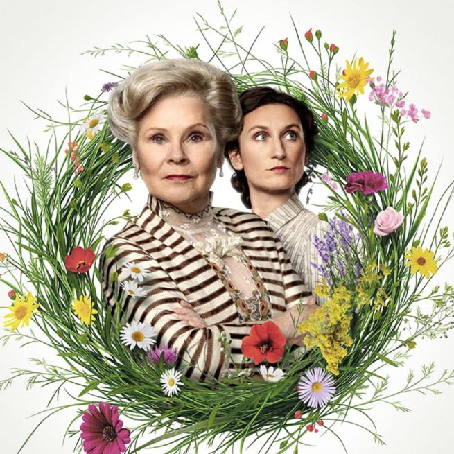 Imelda Staunton stands with arms folded inside a bright floral wreath, as Bessie Carter stands behinds her looking off to the side.