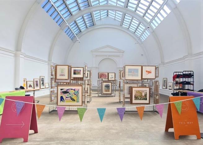 Framed pictures on stands in a large white room with a glazed ceiling, there is colourful bunting and information boards in the foreground