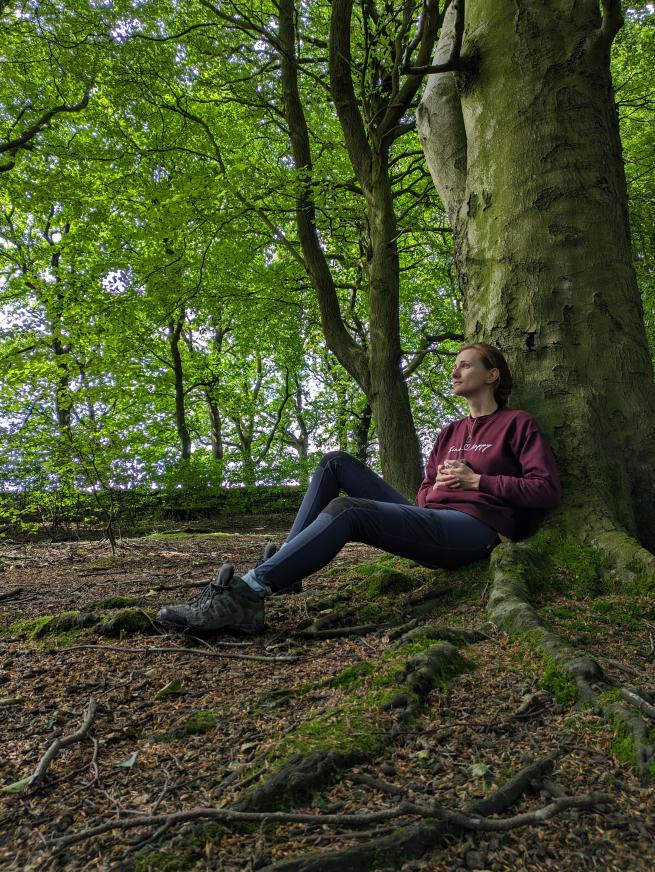 A lady sat at the base of a tree in a woodland holding a mug and looking reflectively 