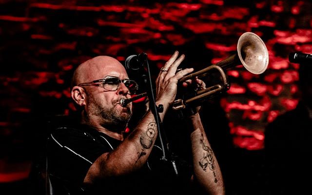 A man (Philip Lassiter) with glasses and tattoos playing a trumpet