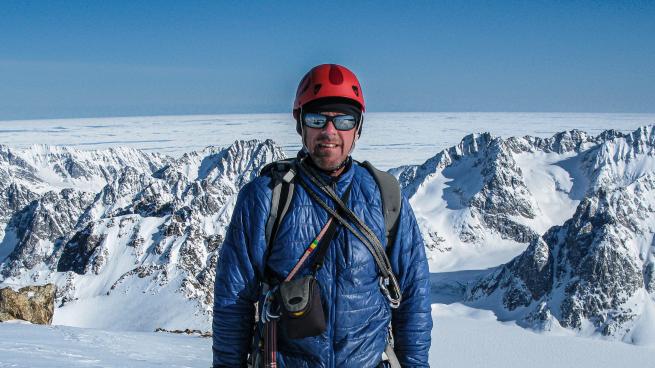 A man in climbing gear stands with a background of snow-covered mountains.