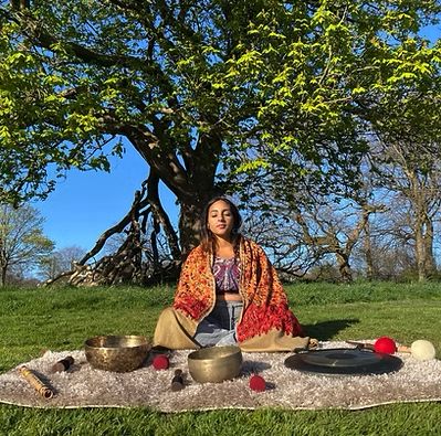 Neeta sat in a field on a blanket with gong, singing bowls and other instruments. Clear warm sky, trees in background. Overall vibe is warm and serene