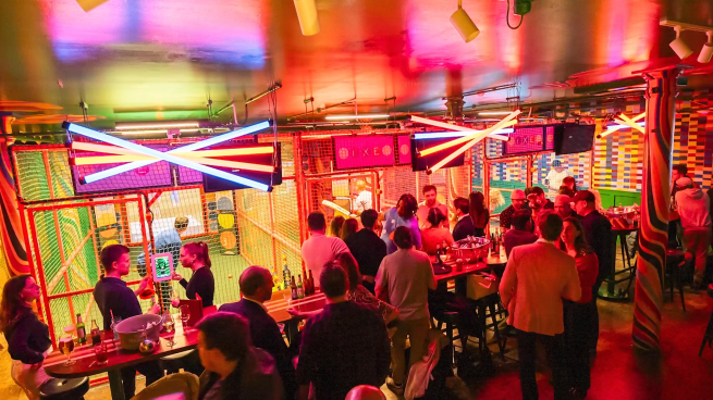 A lively indoor bar scene at Sixes Cricket Club, with groups of people chatting, drinking, and watching others play interactive indoor cricket inside brightly lit batting cages. The venue glows with neon lights and colourful decor, creating a vibrant, energetic atmosphere. Tables are filled with drinks, and the crowd appears relaxed, social, and engaged.