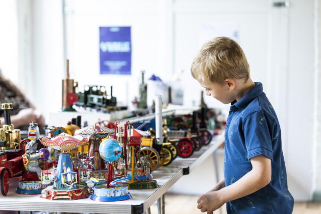A small boy with blonde hair looking at a table full of steam toys