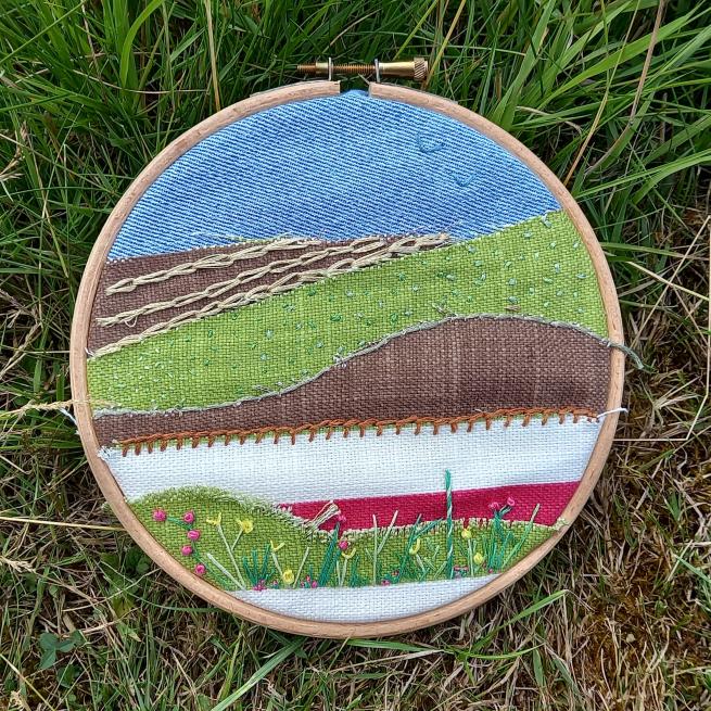 A wooden hoop photographed against a grassy background. The hoop shows a fabric landscape using colours such as blue, brown, green and white 
