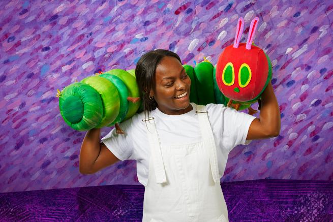 A person stands with The Very Hungry Caterpillar puppet resting across their shoulders, the background is purple.