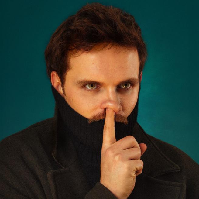 Vittorio Angelone with a turtle neck pulled up his mouth and his finger over where is mouth would be in a 'be quiet' gesture.