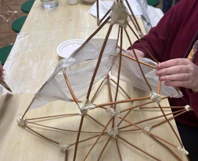 Hands working on a willow sculpture, wrapping material around a geometric structure