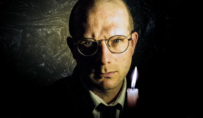 A man holding a candle in a dark room looks intensely into the camera.