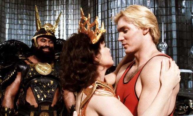 Flash Gordon embraces a woman whilst Brian Blessed looks on at them