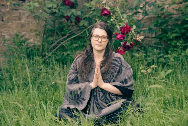 Sophie sat in relaxing clothing, sat cross legged, hands in namaste. Sat in medium length grass 