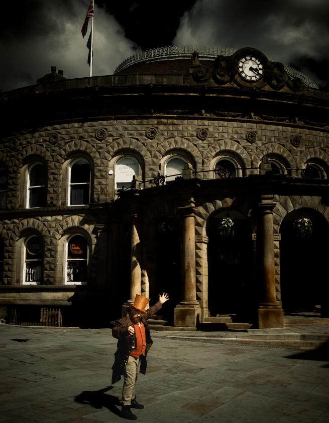 Ghost tour guide, Lord Bartholomew Catch stands in front of Leeds Corn Exchange, welcoming you to his tour under a thundery sky. He wears brown Victorian clothes, a long brown jacket, waistcoat and tweed trousers. He stands with one arm in the air and another holding a lantern. 