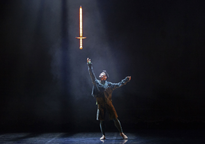On a dark background, a dancer reaches a hand up towards a golden sword which floats above.