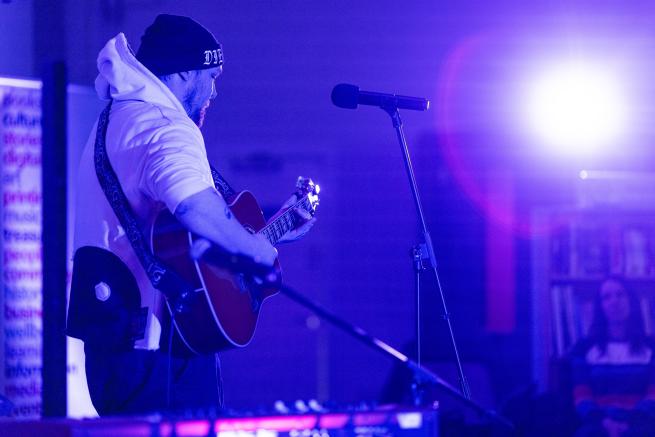 Male in white hoodie Blue lit room playing Guitar singing to a mic with white star burst of light on top right 